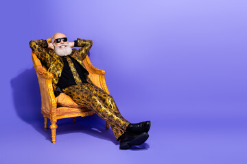 Confident stylish elderly man in a vibrant patterned suit sitting in a golden armchair against a...