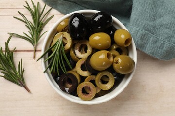 Different delicious marinated olive rings, whole ones and rosemary in bowl on light wooden table, flat lay