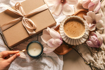 A woman lights a soy candle in a dark glass jar with a matchstick, surrounded by a kraft gift box, blooming magnolia flowers, and a cup of coffee on soft embroidered fabric in warm light