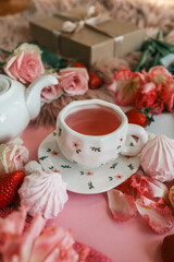 Romantic tea setup with a white teapot, pink tea in a floral cup, candle, roses, tulips, strawberries, and meringues arranged on a pink background with a cozy knitted fabric