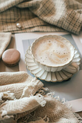 Cup of frothy coffee on a white scalloped saucer, styled with neutral textiles, a macaron, and a fashion magazine. Cozy and elegant aesthetic in soft beige tones
