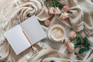 Flat lay of an open planner with a golden pen, cup of frothy coffee, pink roses, macaron, and cozy beige textiles, creating a soft and elegant workspace vibe