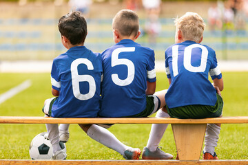 Boys in blue soccer jerseys with white numbers sit on a wooden substitution bench, watching the...