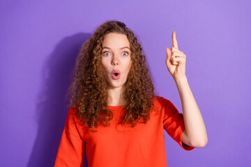 Young woman in casual red outfit expressing surprise and pointing upwards against a vibrant purple...