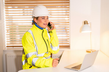 Young woman builder in safety jacket working in office while talking on phone and using laptop...