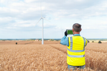 Worker in reflective vest using tablet in wheat field with wind turbine in background in rural area