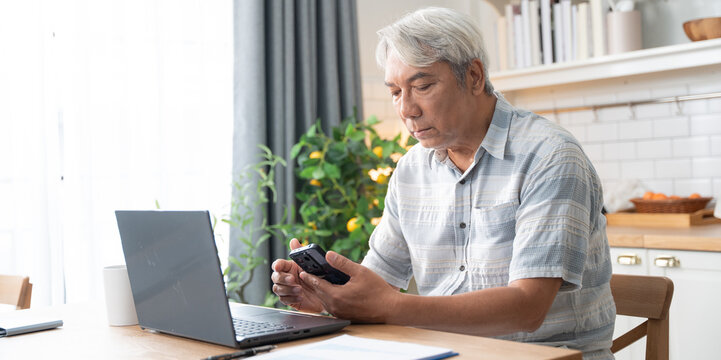Asian senior man checking on mobile phone while working with laptop and document at home. Retired grandfather managing financial payment after retirement - Powered by Adobe