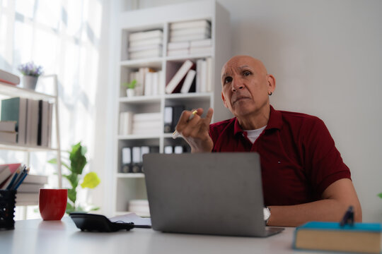 Pensive mature businessman sitting at a desk, holding a pen and gazing thoughtfully away while working on a laptop in a modern office environment, reflecting on strategy and ideas