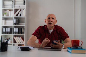 Confident mature businessman having video call using mobile phone sitting at desk in modern office, bookshelf in background, talking and gesturing with hands, explaining something