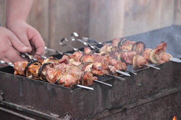Skewers with pieces of meat and vegetables being grilled on a barbecue. Hands turning the skewers over the open flame, showing the cooking process of outdoor grilling.
