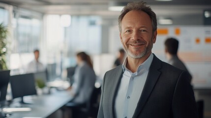 A confident middle-aged businessman smiles in a modern office setting. The environment showcases a collaborative workspace with engaged team members.