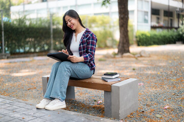 Young college student working on digital tablet, studying outdoors with textbooks, smartphone...