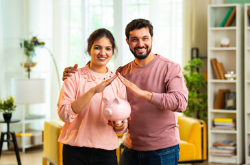 Young Indian couple shows piggy bank with roof hand sign, concept of saving for real estate