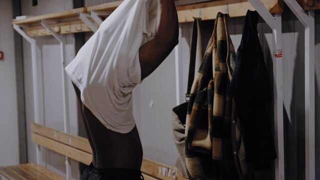 A Black football player removes his casual jersey and puts on his sports uniform in the locker room as part of his pre-game routine.