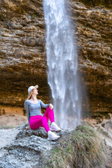 Relaxed young woman in bright sportswear sitting on a rock in front of a scenic waterfall. Outdoor adventure and connection with natur