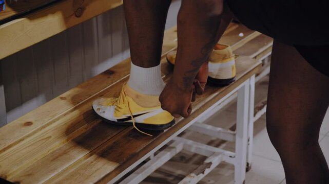 A close-up shot of a Black football player putting on bright yellow boots and tying the laces before the game begins.