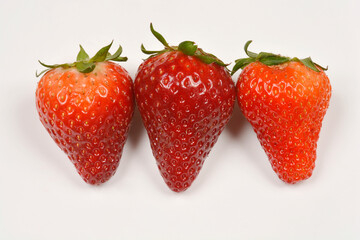 Three fresh strawberries in varying shades of red, each with green leaves, are arranged in a row on a plain light surface