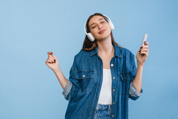 Photo of overjoyed lovely woman dressed denim casual outfit in headphones hand up listen music song using holding mobile phone isolated on blue color background