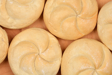 Kaiser rolls are arranged closely together on a light brown wooden surface, viewed from directly...