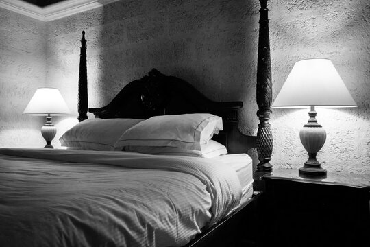 black and white photograph of a double bed with white side of the bed is a lit bedside lamp, the bed has a wooden vintage head board, the light from the two bedside lamps is casting shadows 