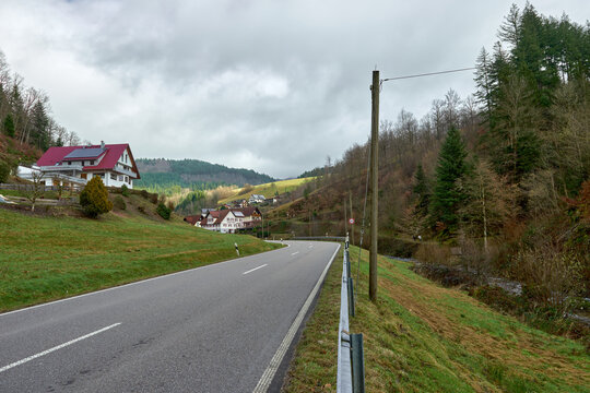 Scenic Mountain Road with Traditional Houses and Red Roofs in Black Forest Valley Landscape. Winding Rural Highway Through Alpine Village with White Buildings and Green Forest Hills Under Cloudy Sky.