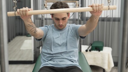 Naklejka premium Man performing seated core exercise on pilates trapeze holding bar to strengthen arms and core muscles at gym. Trainer doing chest press on the cadillac for strengthening chest, shoulder. Habituate.