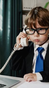 Funny young boy in business suit and glasses making a serious phone call in office