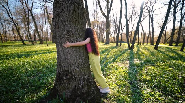 A long-haired brunette in bright clothes hugs a large tree in a sunny park, a moment of unity and grounding with nature to restore mental balance.