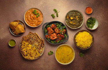 Overhead view of a variety of Indian dishes on a brown table, including curry, tandoori chicken,...
