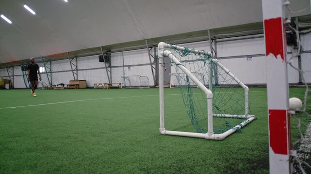A wide shot of a Black football player wearing black sportswear and a pink bandana practicing precision kicks on a small goal and scoring during training.