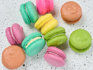 An assortment of colorful macarons on a white surface with dark speckles. The macarons have a smooth, delicate shell and a creamy filling