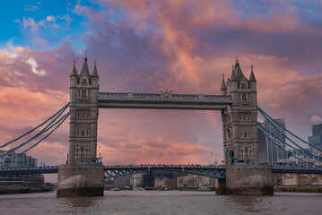 Obraz premium Tower Bridge spans the River Thames in London, featuring intricate towers, suspension cables, and a colorful sky. Modern buildings surround the historic landmark.