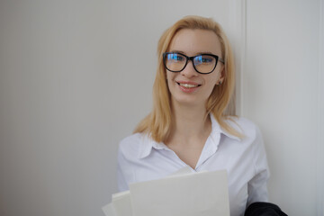 Smiling young woman in glasses holding documents in a bright, modern office setting during the day