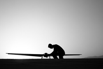 Obraz premium Silhouette of a person inspecting a drone on a desert landscape at sunset.
