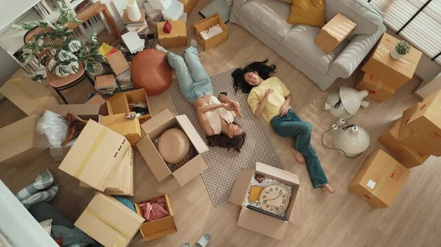 Top down aerial view of lesbian lover lying down at living room with box scattering around and sharing a book to her couple. Asian friend spending time together while sending book to girl. Myrmidon.