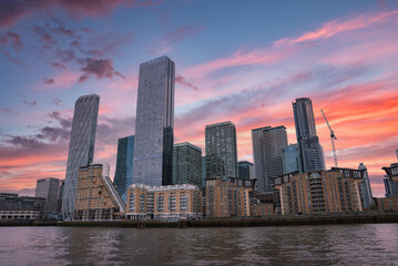 Fototapeta premium View of London's skyline with glass skyscrapers, residential buildings, and a construction crane, reflected in calm river waters at sunset.