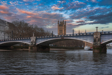 Fototapeta premium Lambeth Bridge spans the River Thames with red and white colors, framed by Victoria Tower, a boat on the water, bare trees, and a sunset sky.