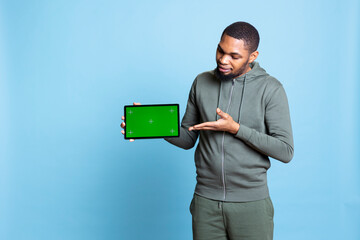 Tech content creator holding isolated green screen tablet doing promotion, talking with audience. Young adult pointing towards chroma key device, doing advertising against studio backdrop.