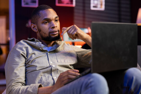 African american adult working remotely from his apartment on laptop, focusing on tasks completion while he lays on a bean bag. Blending productivity and relaxation, browsing the web.