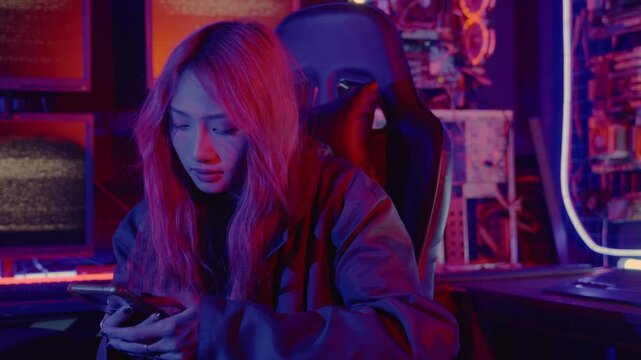 Asian woman in front of a computer monitor display using mobile phone, neon light, Hacker wear black jacket at system control room, Security guard working on multiple monitors together smartphone