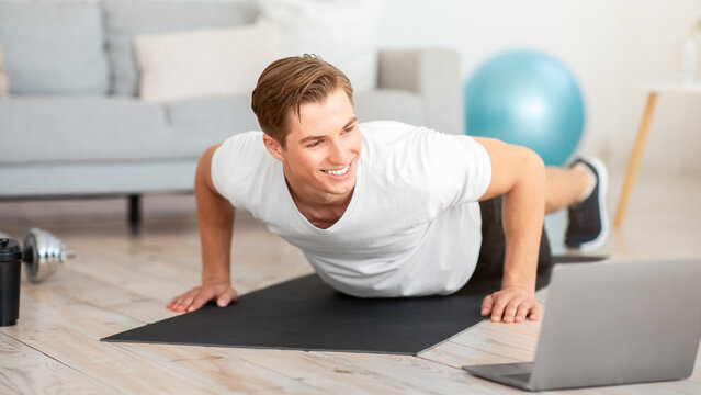 Online training at home remotely. Smiling guy in sportswear does push ups on mat and looks at laptop in living room interior, free space