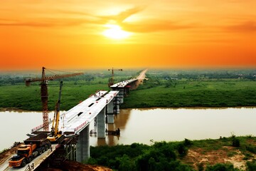 Fototapeta premium Workers carry out construction on a large bridge spanning a river during sunset. Heavy equipment is used, and the surrounding landscape is lush and green.