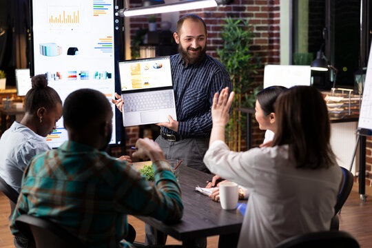 Caucasian businessman reviews brand strategy with startup team, leveraging digital technology for financial planning. The business meeting captures teamwork, innovation and late night productivity.