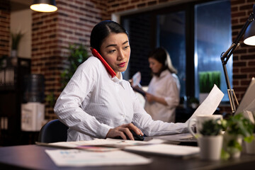 Busy woman juggles multiple tasks, having a phone call, holding a paper with one hand and mouse...