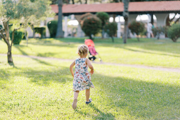 Little girl runs to the stroller across the green lawn. Back view