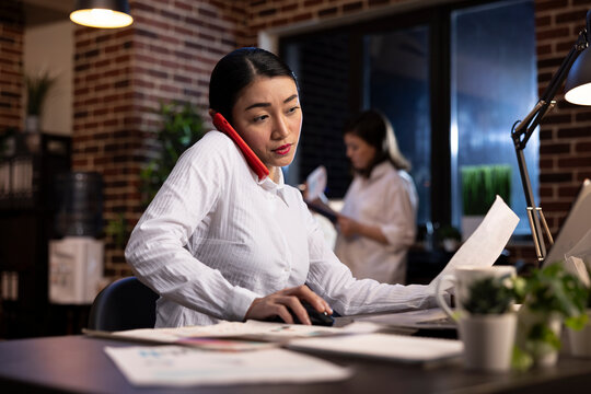 Busy woman juggles multiple tasks, having a phone call, holding a paper with one hand and mouse with the other, all while working on her laptop. Young woman managing late night work responsibilities.