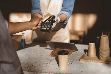 Contactless payment and modern service during social distance and COVID-19. African american customer pays by credit card at online ordering coffee, on terminal, behind bar with waiter, cropped