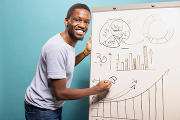 Young black man writing on whiteboard and explaining a chart, standing against a blue studio background. Smiling african american guy holding a pen and using a marker board to showcase a graph.