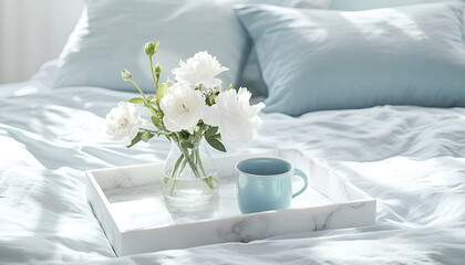 Fototapeta premium Marble white tray holding white flowers in a vase and a blue pastel cup on a bed with pastel blue linens and pillows