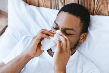 Sick African American Guy Blowing Runny Nose In Paper Tissue Lying In Bed At Home. Ill Black Man...